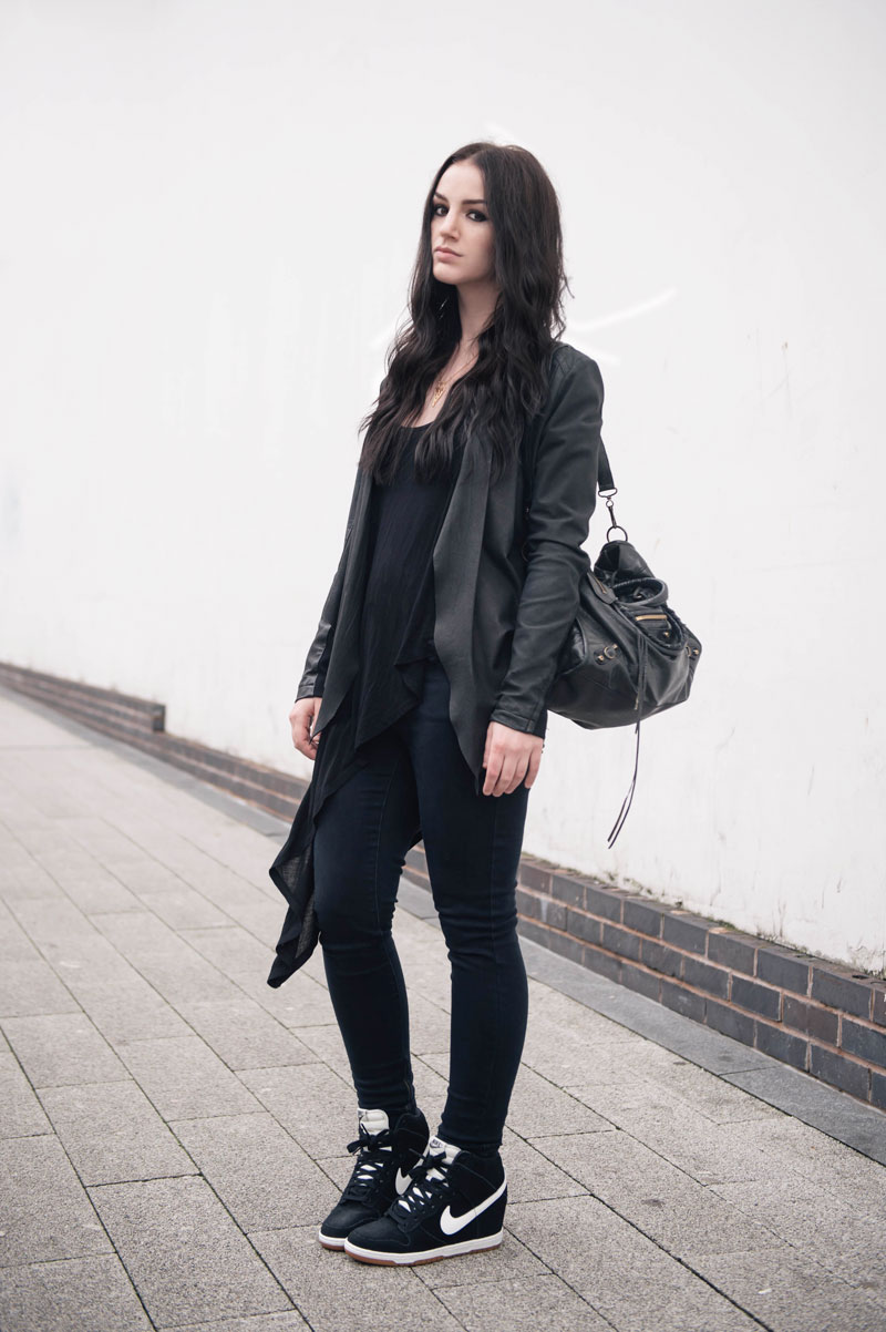 Fashion blogger Stephanie of FAIIINT wearing Barneys Originals draped leather jacket, Topshop skinny jeans & asymmetric top, Nike Sky Hi High Dunk sneakers in black & white, Balenciaga city bag. Casual all black street style outfit.