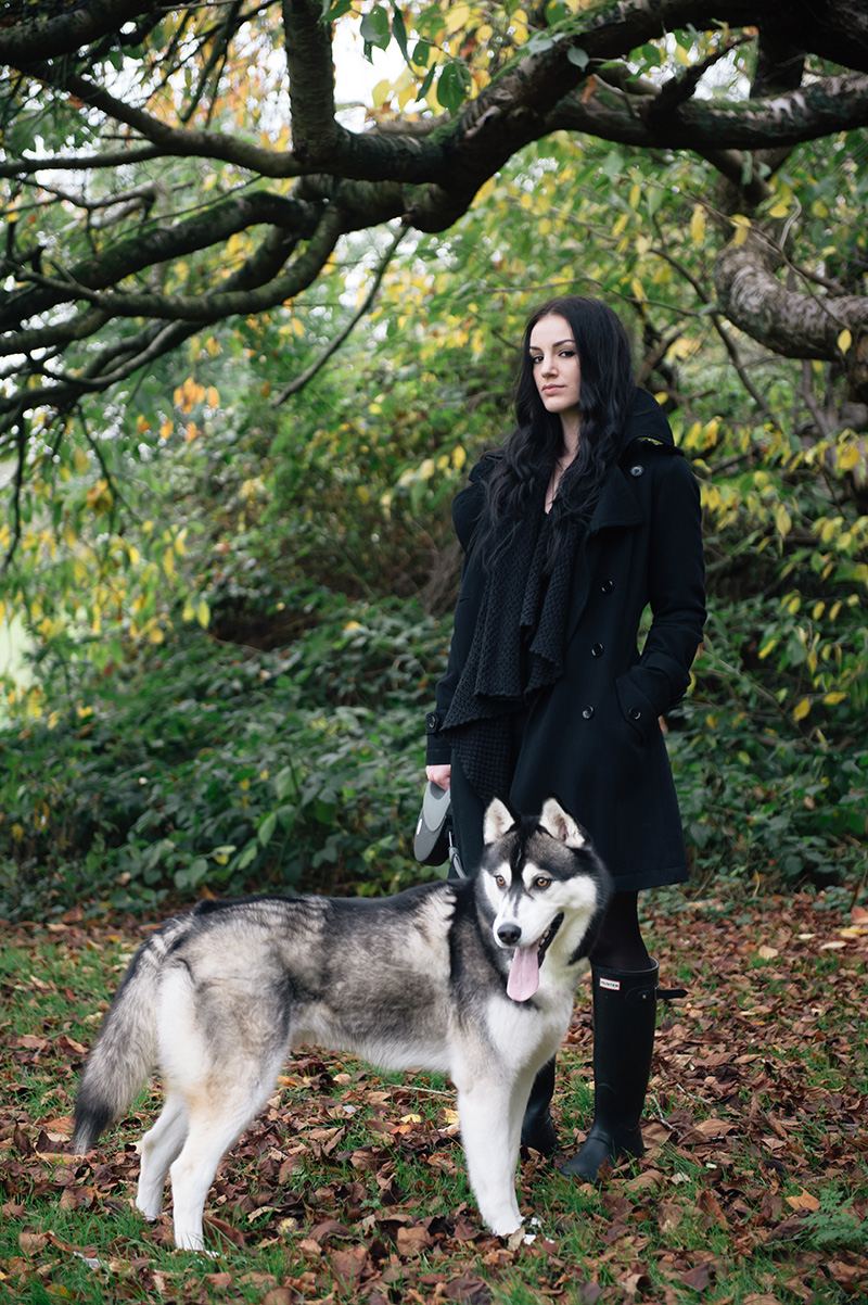 Stephanie Brown of FAIIINT wearing Topshop wool trench coat, ASOS draped chunky cardigan, Hunter wellies wellingtons, all black casual dog walk outfit with Nico siberian husky. Autumn in the park forest leaves on floor, beautiful nature.